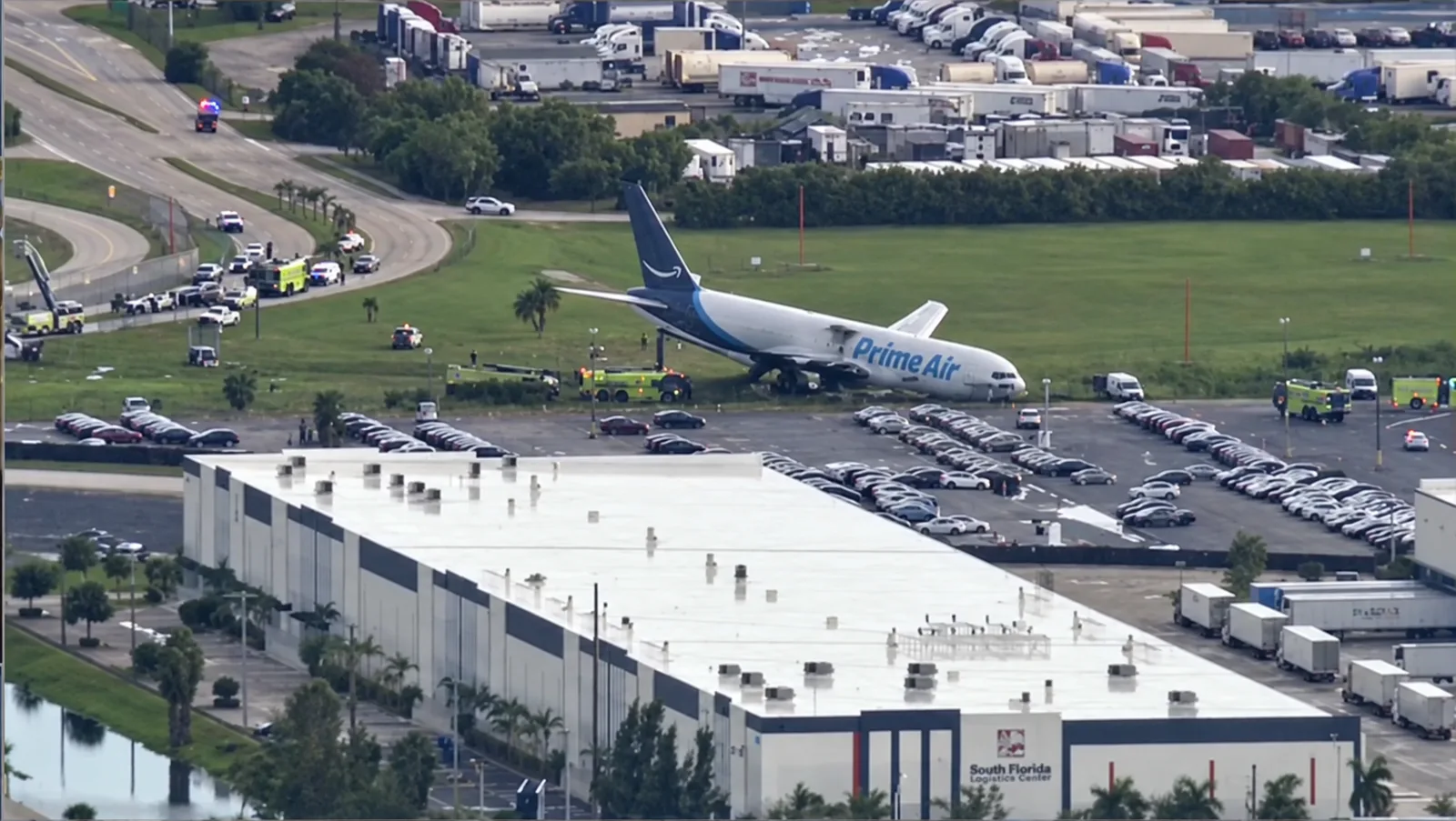 Aerial view of the Amazon Prime Air Boeing 767-300 freighter at rest in a field beyond the runway at Miami International Airport, with fire and rescue units and emergency vehicles surrounding it along NW 42nd Avenue.